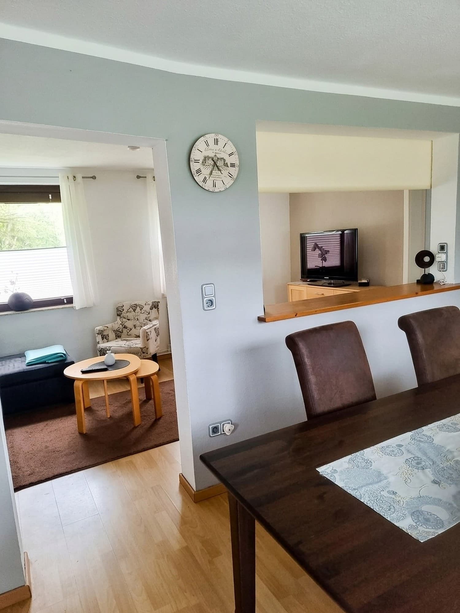 Blick vom Esstisch durch eine offene Tür ins Wohnzimmer, an der Wand eine Uhr, rechts ein Durchblick mit Fernseher