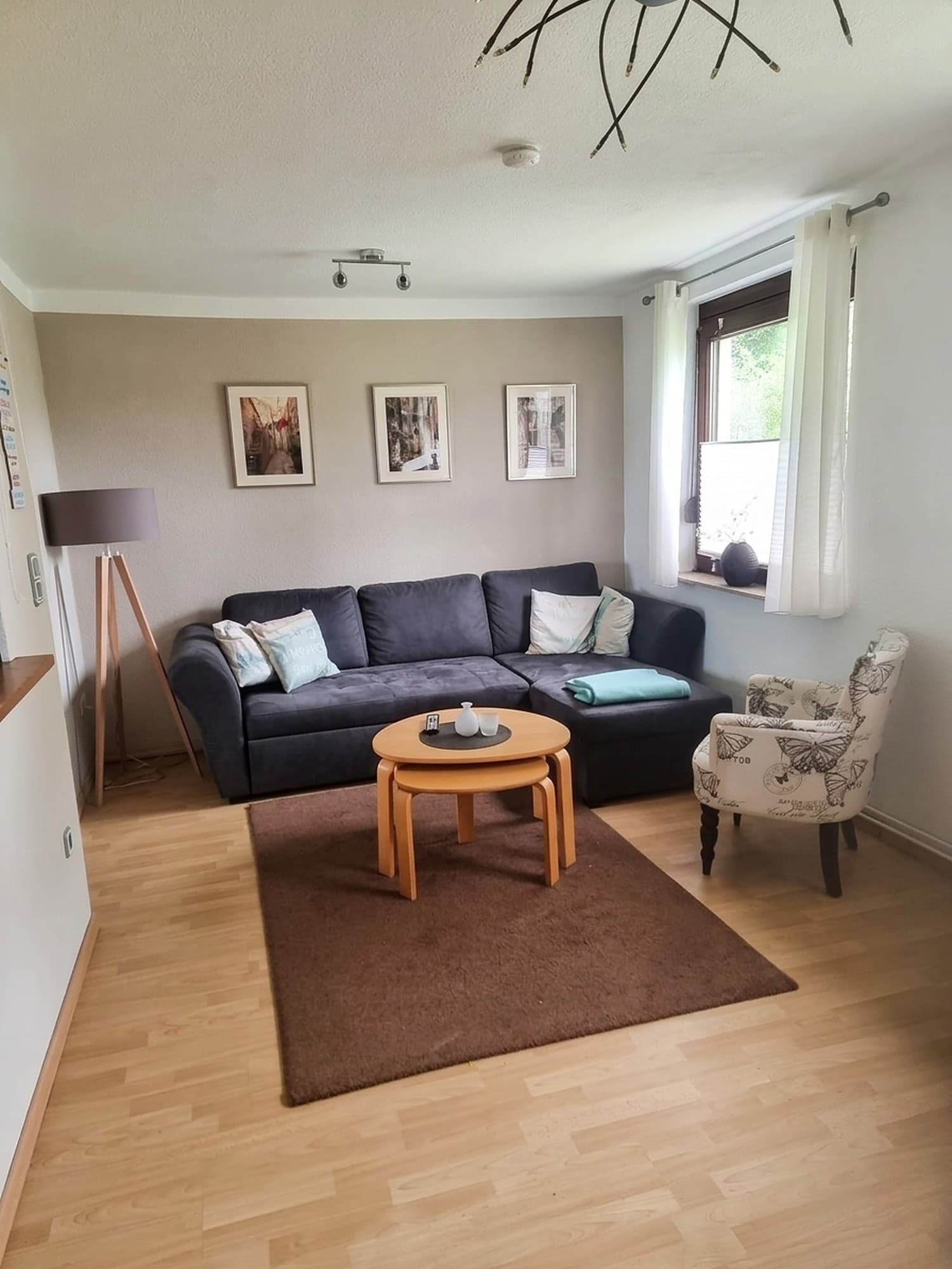 Wohnzimmer mit dunkelgrauem Ecksofa, rundem Couchtisch, Sessel mit Schmetterlingsmuster, Stehlampe und Fenster